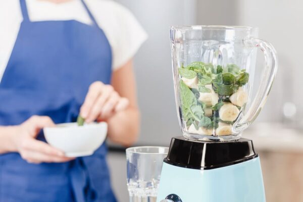 A woman prepares to put more ingredients into a blue blender filled with banana slices and spinach leaves that's sitting on a kitchen counter