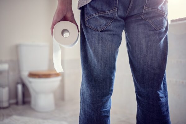 A man in blue jeans and a white shirt stands in front of a toilet holding a roll of toilet paper in his hand