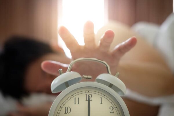 An Asian man laying in bed reaches to turn off an alarm clock