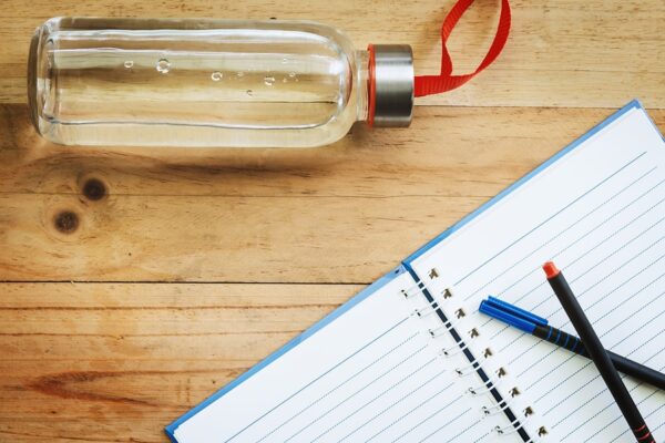 A glass bottle filled with water sit beside a blank notebook and pens on a wooden table